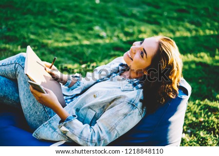 Dreamy successful designer with brunette hair holding notepad for images looking up and thinking on new ideas for drawing cool picture lying down on pouf in park enjoying good weather