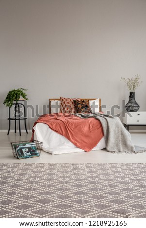 Double bed with bedding, pillows and blanket between bedside table with flower in vase and plant in pot on the small table, real photo with copy space on the wall