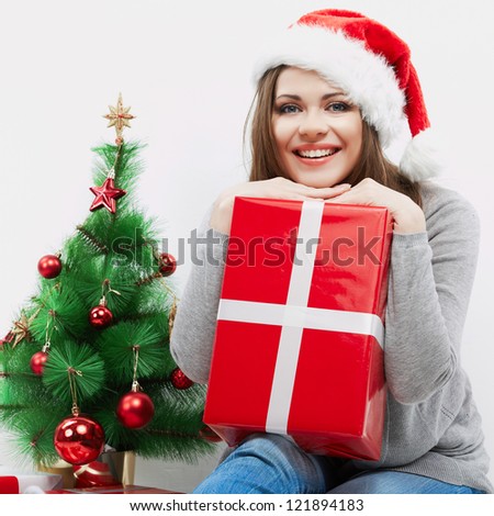Christmas Santa hat isolated woman portrait hold christmas gift beside green  Christmas Tree. Smiling happy girl seat against white wall.