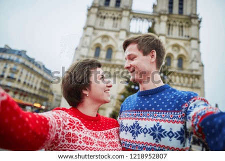 Happy couple in colorful sweaters taking selfie with mobile phone near Notre-Dame cathedral and decorated Christmas tree in Paris, France