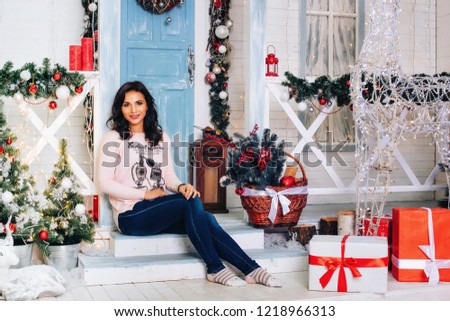 Pretty smiling woman in a pink oversize sweater, jeans and socks on her feet near the festive threshold of the lady. New year's decoration.
