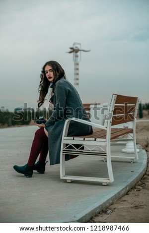A young beautiful girl in a gray coat, gray skirt and burgundy stockings sits on a bench with a vintage camera in her hands.Without retouching