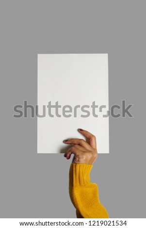 Blank white paper in hand of woman wearing mustard yellow sweater with fingernails painted on grey isolated background for number, letter, words written. (close up, space for text)