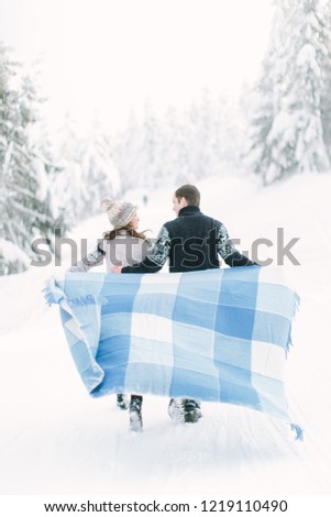 Young couple in love having a walk in winter snowy forest and holding warm wool blanket in hands.
