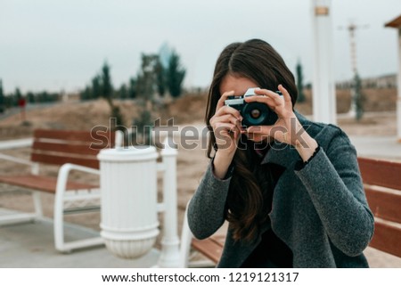 Young beautiful girl in a gray coat, gray skirt and burgundy stockings sits on a bench in the evening and photographs with a vintage camera