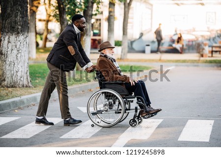 side view of senior disabled man in wheelchair and young african american cuidador riding by street Royalty-Free Stock Photo #1219245898