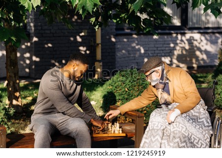 senior disabled man in wheelchair and african american nurse playing chess together on street