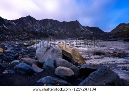 A long exposure photo from above glacial valley. Boulders are in the foreground and an Icelandic glacier is in the background with the sky moving above the scene.