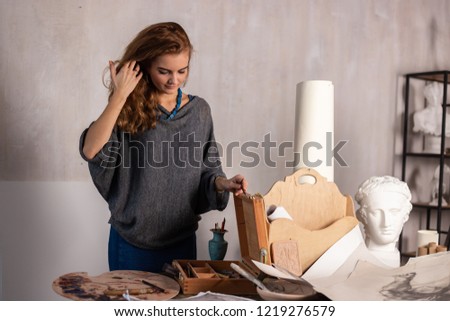 Artist painting on easel in studio. Girl paints landscape with brush. Female painter seen from side. Indoor home interior for handmade crafts.