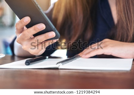 Closeup image of a woman holding and using smart phone with notebook and pen on wooden table