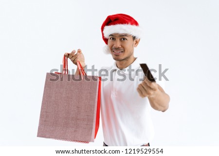 Guy wearing Santa hat, holding shopping bags and paying by credit card. Asian man looking at camera. Christmas shopping and payment concept. Isolated front view on white background.