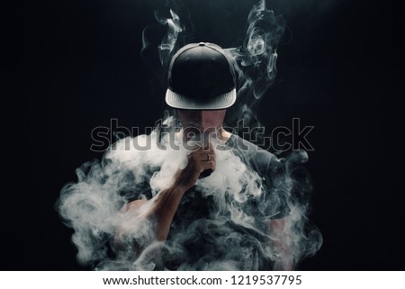 Vape man. Portrait of a handsome young white guy in a modern black cap vaping and letting off puffs of steam from an electronic cigarette  Royalty-Free Stock Photo #1219537795