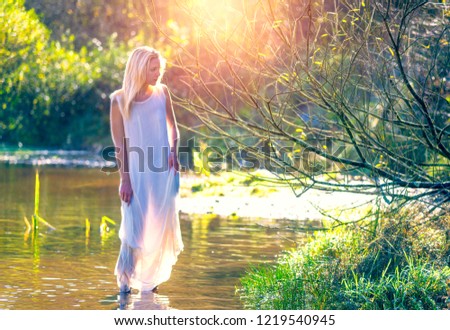 Romantic picture of a spiritual blonde woman in sheer white dress walking barefoot in secluded stream in the countryside with golden sunlight shining through the trees and in her hair.