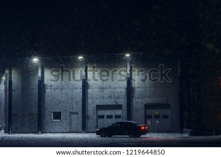 Black car parked near office building in winter evening in strong snow storm. Automobile near auto service in night. Machine near automatic gate for maintenance. Modern architecture in dark night.