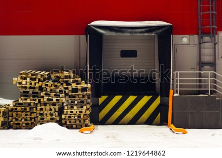 Cargo lift for storage with yellow black diagonal lines warning sign. Wooden pallets on snow. Door for auto loading goods with copy space. Red wall of warehouse in winter. Industrial building close up