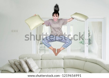 Picture of excited Asian woman holding pillows while jumping on the couch. Shot at home