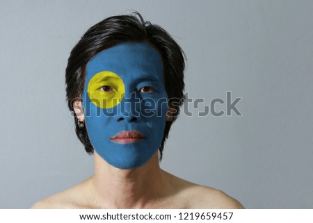 Portrait of a man with the flag of the Palau painted on his face on grey background. A light blue field with the large yellow disk shifted slightly to the hoist-side.
