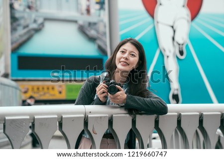 young lady joyfully relaxing on the bridge with camera. girl traveler standing in front the billboard wall in osaka. Travel Japan summer holiday girl enjoying city taking picture.