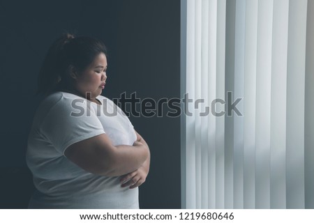 Picture of lonely obese woman looks sad while standing near the window
