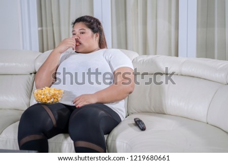 Picture of bored fat woman watching television while eating popcorn on the couch 