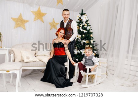 A young family is sitting near a Christmas tree in a white room in New Year's costumes and smiling