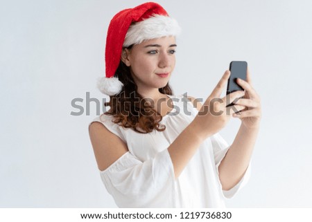 Positive girl in Santa hat having video call, testing camera or mobile app. Pretty young woman using smartphone. Connection concept