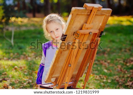 Blond sad little girl drawing on the easel outdoor in summer park. the child paints. Open air activity for school age children concept.