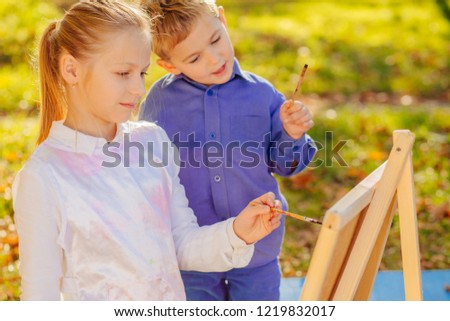Two cute little blond children brother and sister are holding brushes together painting picture with easel outdoor in park. Open air activity for school age children concept.