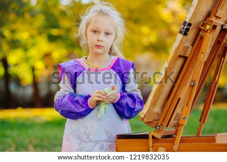 The girl wipes her hands with a napkin while drawing outdoor in fall autumn park. Creative child painting on nature with easel. Activity for kids concept.