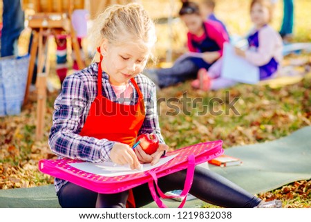 Cute little blonde school girl eating apple and drawing in fall park. Creative child painting on nature. Outdoors activity for school age children concept.