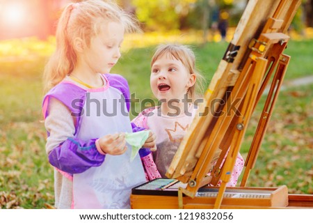 Cute little blond child girl drawing outdoor with other little baby girl in autumn park. Creative child painting on nature with easel. Activity for kids concept.