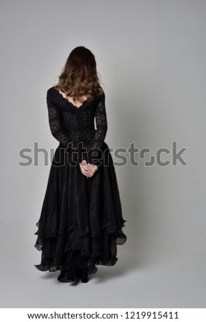 full length portrait of brunette woman wearing long black dress and corset. standing pose with back to the camera, grey studio background.