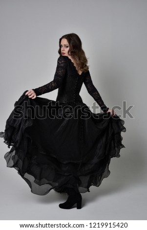 full length portrait of brunette woman wearing long black dress and corset. standing pose with back to the camera, grey studio background.