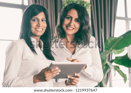 Two happy business ladies using tablet computer in cafe. They are standing and looking at camera with windows in background. Technology concept. Front view.