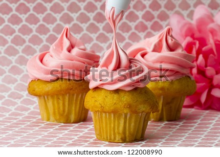 Icing being squeezed onto three cupcakes lying on a pink surface.