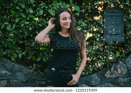 A brunette female with long hair wearing a blank black t-shirt i while standing on wall background on a street. Empty space for text or design.