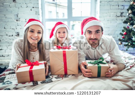 Christmas, holidays, family and people concept - happy mother, father and little girl in santa helper hats with gift boxes over living room and christmas tree background