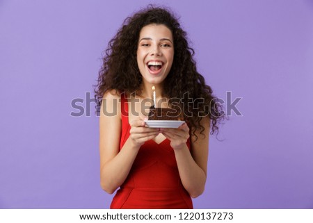 Image of happy woman 20s wearing red dress holding piece of birthday cake with candle isolated over violet background