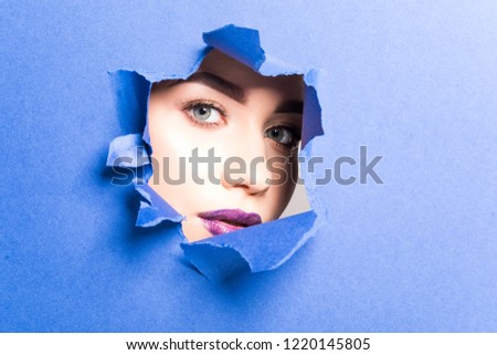 The face of a young beautiful girl with a bright make-up and puffy purple lips peers into a hole in violet paper.
