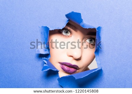 The face of young beautiful girl with a bright make-up and puffy purple lips peers into a hole in purple paper.