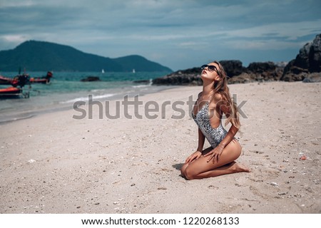 Young fit woman in a leopard swimsuit and black sunglasses sitting on the wet sand beach with out of focus boat in background. Karon beach. Phuket.