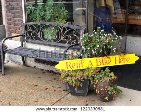 Sign outside local small town store indicating that bikes are available for rent - next to potted plants and wrought iron bench