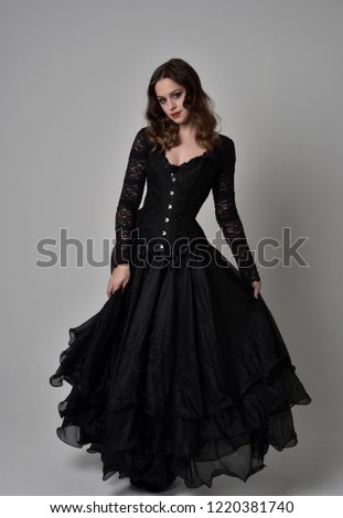 full length portrait of brunette girl wearing long black lace gown. standing pose on grey studio background.
