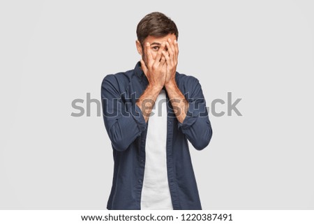 Handsome man with dark hair, bristle, hides face with hands, peeks through fingers, wears fashionable shirt, feels shy, isolated over white background. Curious guy cannot to look at present. Royalty-Free Stock Photo #1220387491