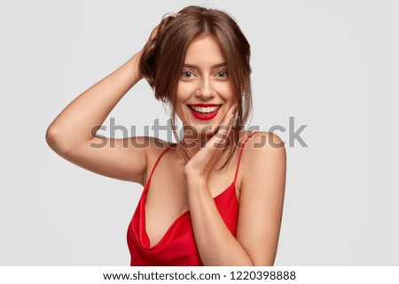 Headshot of beautiful female model has healthy skin, positive expression, poses for fashion magazine, collects hair, wears red outfit and lipstick, isolated over white background. Beauty concept