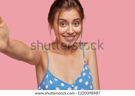 Smiling woman with joyful expression, stretches hand for making selfie, has healthy soft skin, pleasant appearance, models against pink background. Caucasian female takes picture of herself.