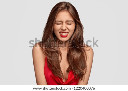 Aggressive young lady clenches teeth with annoyed expression, doesnt like something, closes eyes from despair, wears red lipstick, expresses negative feelings, stands in studio against white wall