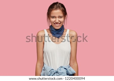 Positive shy woman has tender smile, bites lower lip, recieves intriguing suggestion, dressed in casual white t shirt, has shirt wrapped around waist, poses against pink background. Emotions concept