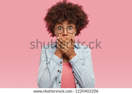 Shot of pleasant looking young ethnic female covers mouth with both palms, tries not spread rumors, being shocked with gossiping, stares through round spectacles, stands against pink background.