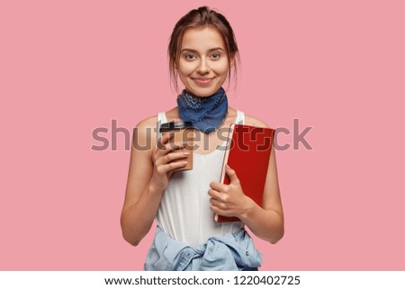 Picture of pleasant looking young schoolgirl dressed in fashionable outfit, drinks takeaway coffee, reads something from red textbook, likes have talk together with groupmates during break at college